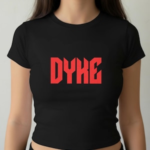 May include: Black crop top with the word "DYKE" in bold, red, block letters across the chest. The shirt has short sleeves and a crew neck. The text is centered and prominently displayed.