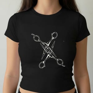 May include: Black t-shirt featuring a white graphic of crossed scissors. The design is centered on the front of the shirt. The shirt has a crew neck and short sleeves.