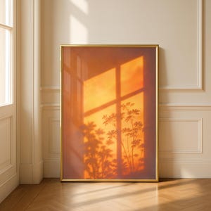 May include: A vertical rectangular wall art piece in a thin gold frame. The print displays a warm, orange-toned sunset scene featuring the distinct silhouette of a leafy plant cast against a window pane, creating a soft, glowing light effect.