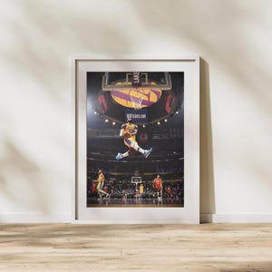 May include: Framed print of a basketball player mid-air, about to dunk the ball. The player is wearing a yellow and purple uniform. The background shows the basketball court, the audience, and the Lakers logo.