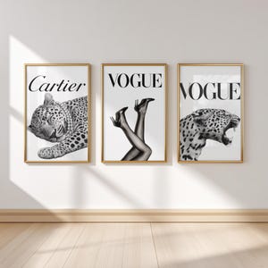May include: Three framed art prints with gold frames. The prints feature a black and white leopard, the word "Cartier", the word "Vogue" with legs in heels, and a roaring leopard. The prints are displayed on a white wall.