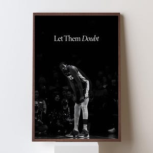 以下が含まれることがあります： バスケットボール選手が身を乗り出し、白い文字で「Let Them Doubt」と書かれた白黒の額入りプリント。画像は、背景に人々の群衆がいる暗い背景に対して設定されています。