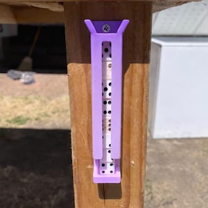 May include: A light purple plastic vertical holder mounted to a wooden post with a single screw. The holder contains a stack of white dominoes with black pips, visible through the open front channel of the rectangular storage unit.