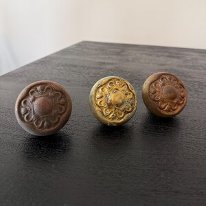 May include: Three round metal cabinet knobs with a raised floral relief pattern. The knob on the left has a dark brown patina, the centre knob is bright gold with blue oxidation, and the right knob has a muted bronze finish with dark accents.
