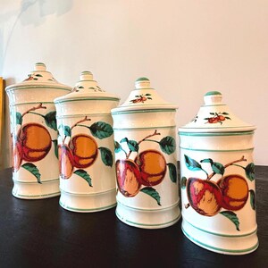 May include: Four matching ceramic canisters with lids. The canisters are off-white with a green trim and feature a hand-painted design of peaches and green leaves. The lids have a small knob and a peach design. The canisters are on a dark surface.