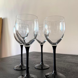 May include: Four clear wine glasses with black stems and bases are arranged on a dark surface. The glasses have a classic, elegant design, perfect for serving wine. The stems and bases are a glossy black, contrasting with the clear bowls.