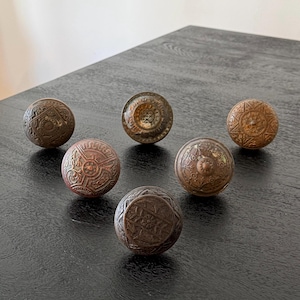 May include: A collection of six round, metallic door knobs featuring intricate, embossed geometric and floral patterns. Each knob displays a weathered, aged bronze finish with varying levels of patina, showcasing detailed textures and relief designs.