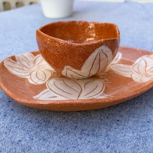 Peut inclure: Un ensemble en céramique fait main composé d'un petit bol rond orange moucheté au bord irrégulier et d'une assiette ovale assortie. Les deux pièces présentent des motifs de pétales de fleurs blancs peints à la main sur l'argile terre cuite.