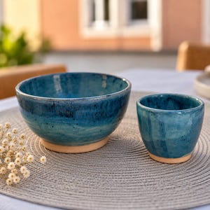 Könnte beinhalten: Eine Keramikschale und ein passender Becher mit einer glänzenden, nuancierten blauen Glasur mit dunklen Sprenkeln und einem glatten, unglasierten beigen Boden. Das Set ist auf einem runden grauen Web-Platzset neben einem Zweig weißer Trockenblumen arrangiert.