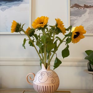 Può includere: Un vaso in ceramica rosa chiaro con corpo rotondo, un singolo manico curvo e un collo stretto. La parte superiore presenta motivi a spirale disegnati a mano in marrone scuro, mentre la sezione inferiore ha dettagli a coste verticali. Contiene diversi girasoli giallo brillante.