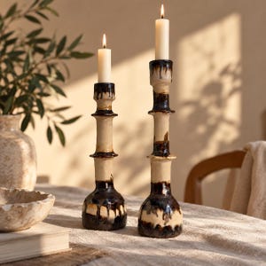 Può includere: Due portacandele in ceramica di diverse altezze, ciascuno con una base color crema e dettagli in smalto marrone scuro colante. Entrambi sostengono una candela bianca accesa, caratterizzati da un design cilindrico a gradini con base larga.