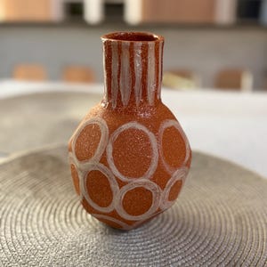 Op de afbeelding: Een terracotta-kleurige keramische vaas met een gespikkelde textuur, voorzien van een rond lichaam en een smalle hals. Het oppervlak is versierd met handgeschilderde witte cirkelvormige contouren op het lichaam en verticale witte strepen op de hals.