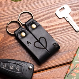 May include: Black leather keychains with gold-colored hardware, featuring the letters "A" and "B" with a heart design. A car key, a house key, and a patterned notebook are also visible on a wooden surface.