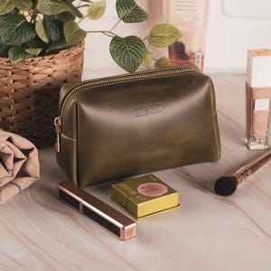 May include: An olive green leather cosmetic bag with the initials "KND" embossed on the front. The bag has a gold zipper and is surrounded by makeup products, a makeup brush, and a small potted plant. A rolled-up towel is also present.