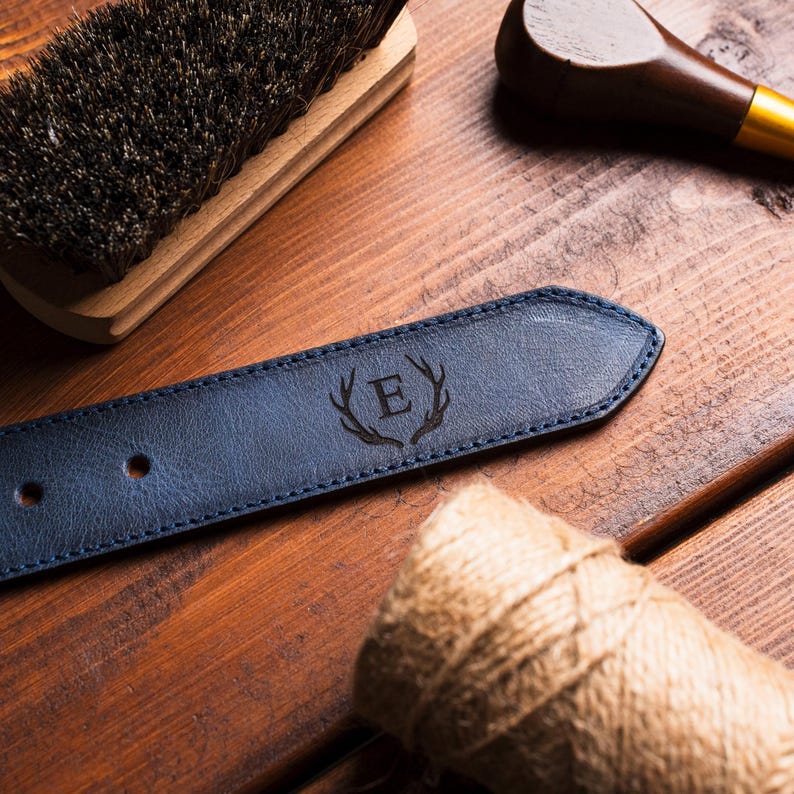 Puede incluir: Primer plano de un cintur&oacute;n de cuero azul marino con la letra "E" y un dise&ntilde;o de cornamenta grabado en la punta. El cintur&oacute;n est&aacute; sobre una superficie de madera con un cepillo, una herramienta de madera y un rollo de cuerda.