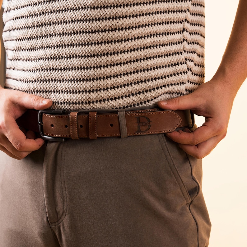 Puede incluir: Un cintur&oacute;n de cuero marr&oacute;n con hebilla de metal oscuro y logotipo estampado. El cintur&oacute;n se usa con pantalones marrones y una camisa a rayas. El cintur&oacute;n tiene unos 3,8 cm de ancho.