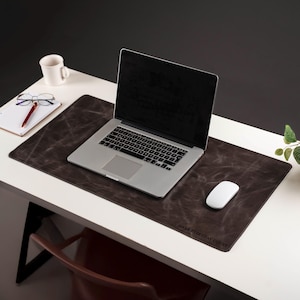 May include: A laptop sits on a dark brown desk mat with the text "MAKE EVERYDAY COUNT". A white mouse, coffee mug, notepad, and glasses are also on the white desk. The background is dark.