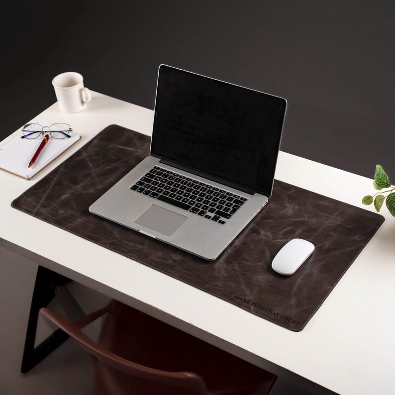 Może przedstawiać: Laptop leży na ciemnobrązowej podkładce pod biurko z napisem "MAKE EVERYDAY COUNT". Biała mysz, kubek, notatnik i okulary r&oacute;wnież znajdują się na białym biurku.