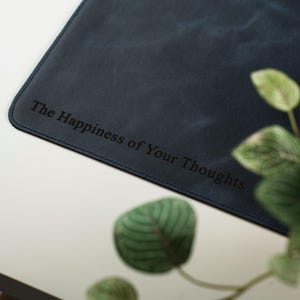 Może przedstawiać: Granatowa sk&oacute;rzana podkładka na biurko z wygrawerowanym napisem "The Happiness of Your Thoughts". Podkładka ma zaokrąglone rogi i szytą krawędź. Zielone liście są widoczne w tle.