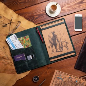 May include: A dark green leather travel organizer with a notepad featuring an elephant sketch, a red pen, and pockets holding a passport, tickets, and a patterned card. A white smartphone and a cup of coffee sit nearby.