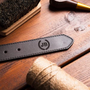 Puede incluir: Primer plano de un cintur&oacute;n de cuero marr&oacute;n oscuro con un borde cosido. El cintur&oacute;n presenta un monograma "JB" en relieve dentro de una corona de laurel. Tambi&eacute;n se ven un cepillo de madera, una pipa marr&oacute;n y un rollo de cuerda.