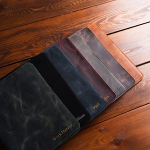 May include: A selection of leather portfolios in shades of green, black, navy, burgundy, grey, and brown. Each portfolio displays a distinct texture and personalised text. The items are arranged on a wooden surface.