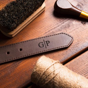 Puede incluir: Primer plano de un cintur&oacute;n de cuero marr&oacute;n con las iniciales "G|P" grabadas en el extremo. El cintur&oacute;n est&aacute; sobre una superficie de madera, con un cepillo, una herramienta de madera y una madeja de cuerda en el fondo.