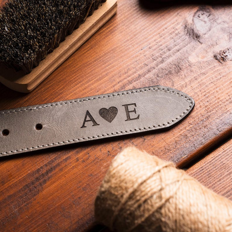 Puede incluir: Un cintur&oacute;n de cuero gris con las iniciales "A" y "E" separadas por un s&iacute;mbolo de coraz&oacute;n. El cintur&oacute;n est&aacute; sobre una superficie de madera con un cepillo y un rollo de hilo en el fondo. El cintur&oacute;n tiene un borde cosido.