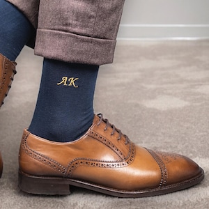 Puede incluir: Zapatos Oxford de cuero marrón con detalles de brogue, combinados con calcetines azul marino bordados con iniciales doradas. Los calcetines son visibles por encima de los zapatos, y la persona lleva pantalones marrones.