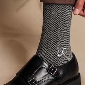 Puede incluir: Zapatos negros de cuero con doble hebilla y suela gruesa. Los zapatos se combinan con calcetines grises de espiga bordados con las letras "CC" en blanco. La persona lleva pantalones marrones.