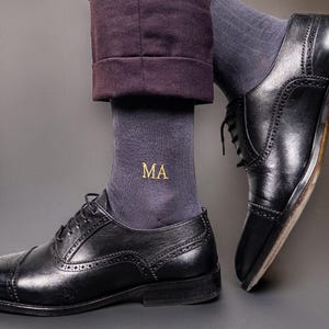 Pode incluir: Sapatos Oxford de couro preto com costura decorativa e design com atacadores. Os sapatos s&atilde;o combinados com meias cinza escuro bordadas com as letras douradas "MA". A pessoa est&aacute; usando cal&ccedil;as roxas escuras.