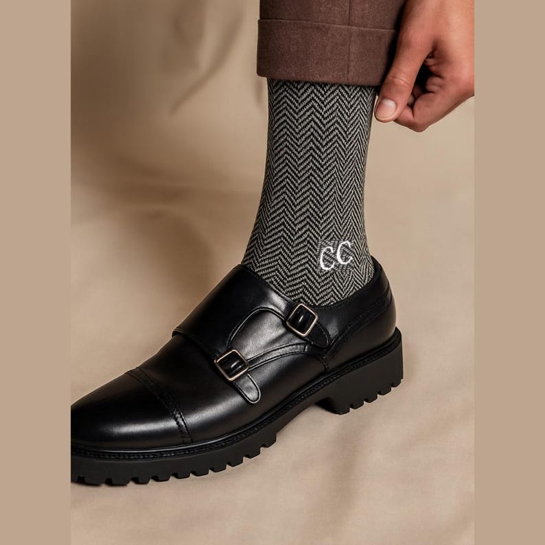 K&ouml;nnte beinhalten: Schwarze Doppelriemenschuhe aus Leder mit dicker Sohle. Die Schuhe werden mit grauen Fischgr&auml;ten-Socken kombiniert, die mit den Buchstaben "CC" bestickt sind. Die Person tr&auml;gt eine braune Hose.