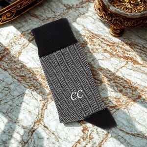 K&ouml;nnte beinhalten: Schwarz-grau gemusterte Socken mit den Initialen "CC" in Wei&szlig; gestickt. Die Socken haben ein Fischgr&auml;tmuster auf dem Hauptteil und einen einfarbigen schwarzen Bund und Zehen. Die Socken werden auf einer Marmoroberfl&auml;che pr&auml;sentiert.
