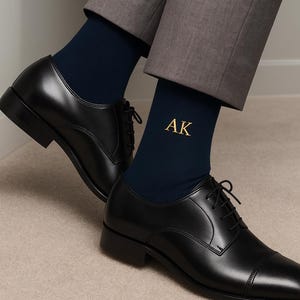 Pode incluir: Meias azul marinho com as iniciais "AK" bordadas a ouro s&atilde;o vis&iacute;veis acima de sapatos de vestir de couro preto. Os sapatos t&ecirc;m um design cl&aacute;ssico com biqueira e s&atilde;o amarrados. Cal&ccedil;as cinzentas.