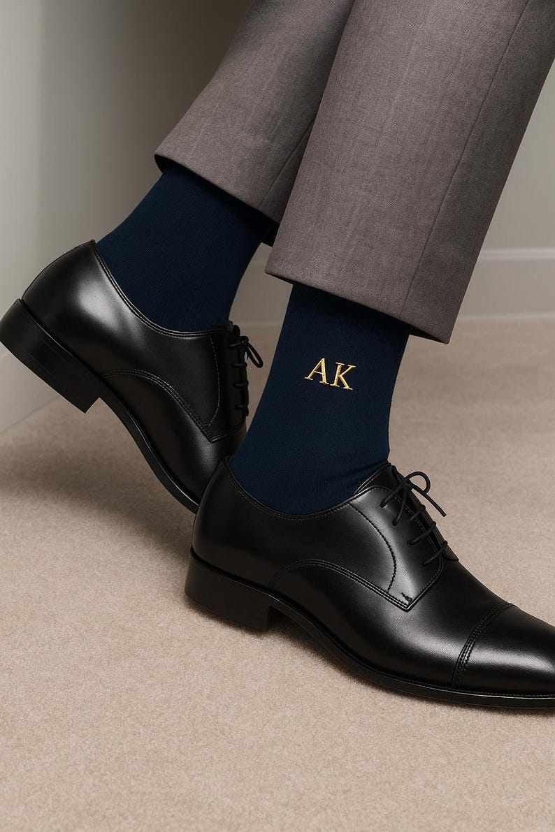 Pode incluir: Meias azul marinho com as iniciais "AK" bordadas a ouro s&atilde;o vis&iacute;veis acima de sapatos de vestir de couro preto. Os sapatos t&ecirc;m um design cl&aacute;ssico com biqueira e s&atilde;o amarrados. Cal&ccedil;as cinzentas.
