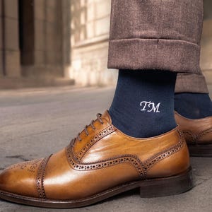 Pode incluir: Sapatos de couro castanho com detalhes brogue e meias azul marinho. As meias s&atilde;o bordadas com as iniciais "TM" em branco. A pessoa est&aacute; usando cal&ccedil;as castanhas.