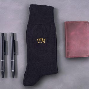 Pode incluir: Um conjunto de acess&oacute;rios, incluindo duas canetas pretas, um par de meias caneladas azul marinho com iniciais douradas bordadas e uma carteira de couro bord&ocirc;. Os itens est&atilde;o dispostos sobre uma superf&iacute;cie cinza.