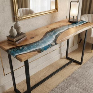 Può includere: Tavolo consolle in legno con un design a fiume in resina epossidica blu e bianca. Il tavolo ha una finitura a venature naturali e gambe in metallo nero. Vasi decorativi e libri sono sul tavolo.
