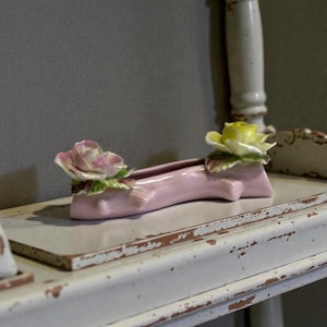 May include: A pink ceramic shoe-shaped vase with a pink and yellow rose on each end. The vase is on a distressed white shelf. The roses have green leaves. The vase is a decorative item.