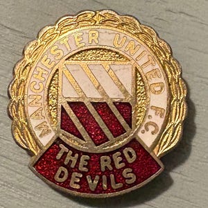 May include: A gold-toned Manchester United F.C. pin with a red and white crest. The pin features the text "MANCHESTER UNITED F.C." and "THE RED DEVILS" in gold lettering. The crest has a red, glittery background.