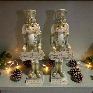 May include: Two silver and gold Christmas nutcracker figures, each holding a sign saying "Merry Christmas". The nutcrackers have white hair and beards, and stand on grey bases. Pine cones and fairy lights are arranged in the background.