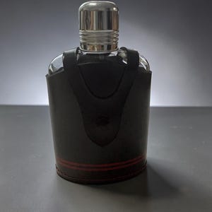 May include: A clear glass bottle with a silver-coloured metal cap, encased in a dark brown leather sleeve. The leather sleeve features a vertical strap design and two thin horizontal red lines near the bottom, secured with visible stitching along the base.