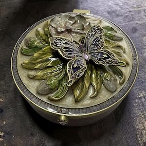 Puede incluir: Una caja decorativa redonda con una mariposa en relieve de alas moradas y doradas sobre un lecho de hojas verdes y una flor grande. La tapa es de color crema con borde dorado y un pequeño cristal transparente en el mecanismo de cierre.