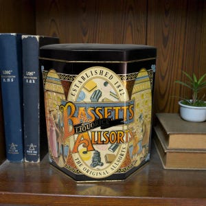 May include: A vintage Bassett's Liquorice Allsorts tin, black and gold, with a hexagonal shape and colorful graphics. The tin is on a wooden shelf with books and a small potted plant.