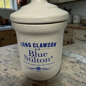 May include: A cream-colored ceramic crock with a matching lid. The front features blue text reading LONG CLAWSON Blue Stilton, with the additional text Stilton makers for over 100 years and a small circular logo at the bottom center.