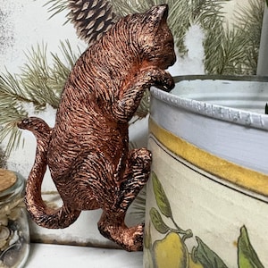 May include: A bronze cat ornament climbing on a container. The cat has detailed fur and is reaching up. The container is white with a yellow border and a botanical illustration of lemons and leaves.