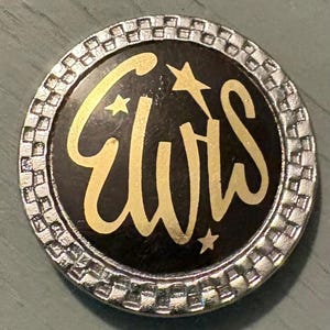 May include: A round, silver-toned pin with the name "ELVIS" in gold script, accented with stars. The center is black, and the outer edge has a textured, checkered pattern.