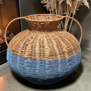 May include: A large, two-tone wicker basket with a round base and two handles. The bottom half is painted blue, while the top half is natural wicker. The basket has a wide opening and is suitable for storage or decoration.