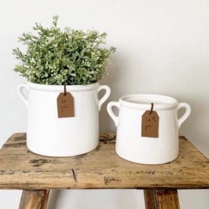 May include: Two white ceramic plant pots with curved handles on a wooden surface. The larger pot contains greenery. Each pot has a brown tag that reads "for you". The pots are a decorative home accent.