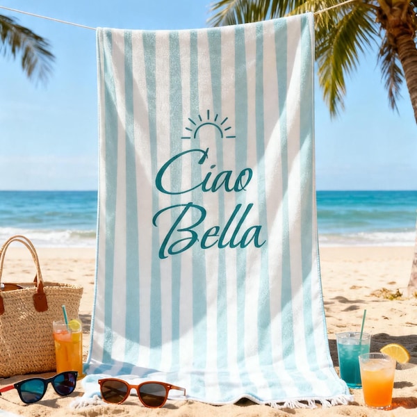Ciao Bella striped beach towel in teal and white, hanging between palm trees at the beach with sunglasses and a straw bag